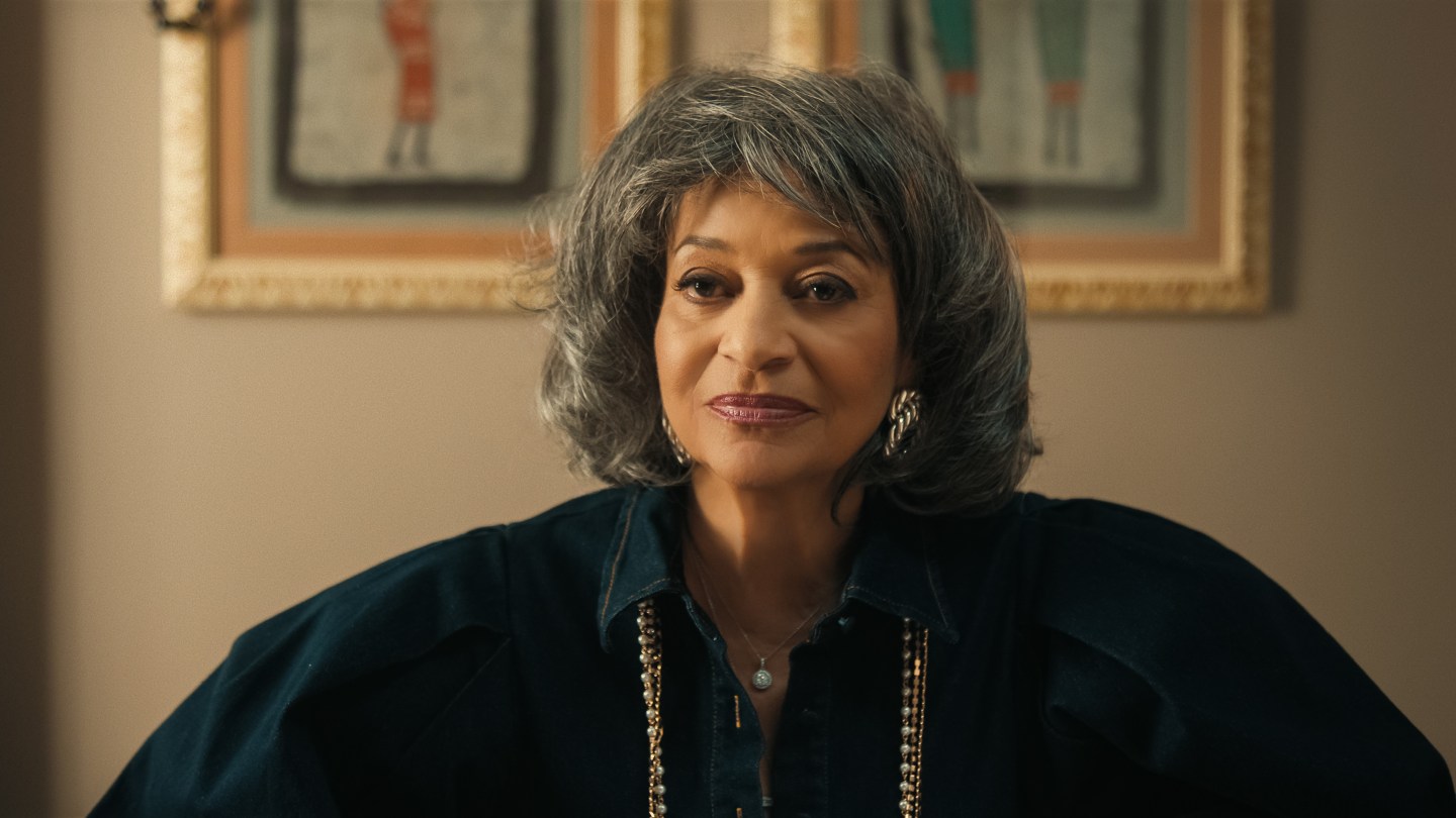"Woman with gray hair wearing a dark blouse and gold necklace, sitting indoors in front of framed artwork on a beige wall, softly lit room."